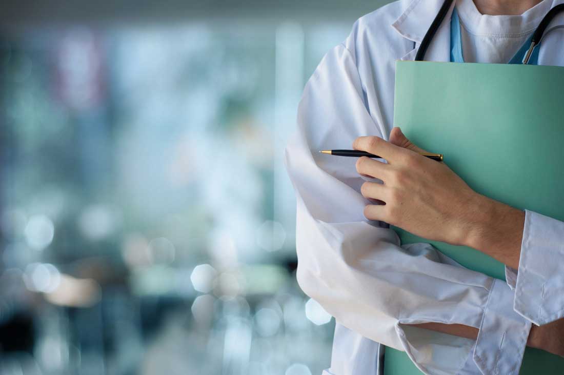 Photo of a doctor from neck to waist in white medical gear holding a chart to their chest